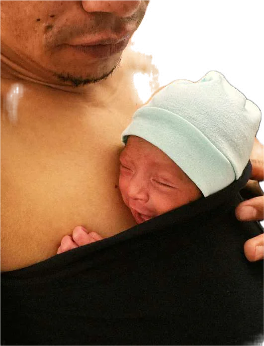 Papá cargando a su bebé con gorro dentro de una faja negra, piel con piel